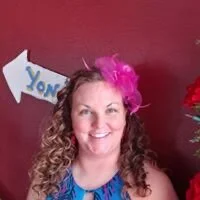 Woman with curly hair wearing a pink feather accessory and smiling, standing in front of a maroon wall with a partially visible 'YON' sign.