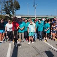 A group of people standing outdoors on a paved area with a bridge in the background, along with one dog.