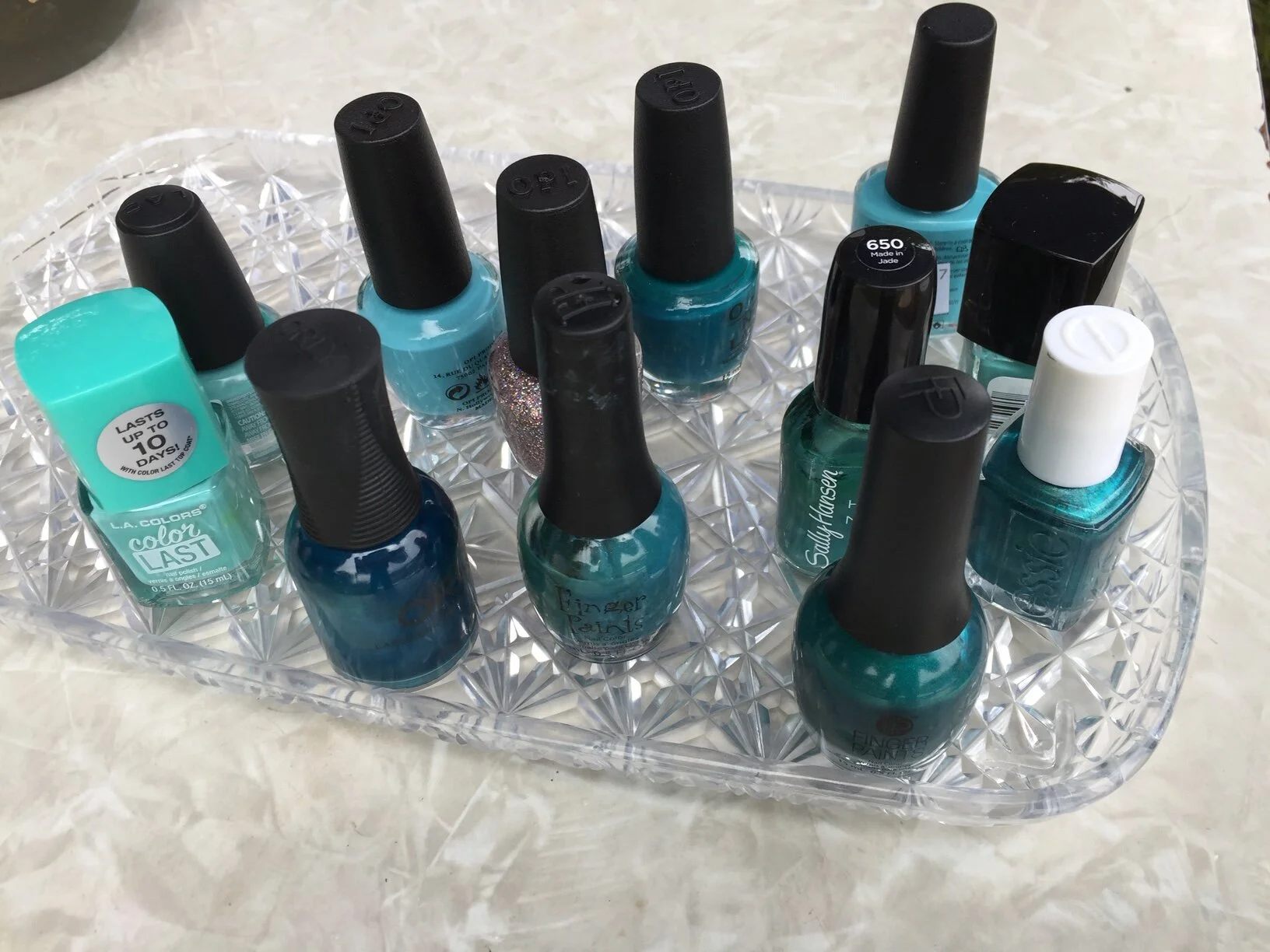 A clear plastic tray holding multiple bottles of blue, teal, and turquoise nail polish, some with black caps and some with white caps, placed on a marble surface.