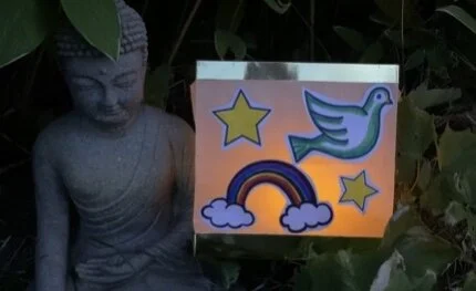 A stone statue of Buddha next to a colorful sign with stars, a rainbow, and a bird, outdoors among green leaves.
