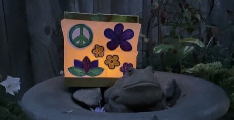 A frog resting on a stone surface next to a colorful sign with peace sign and flower illustrations.