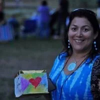 Woman smiling outdoors, holding a colorful painted canvas with a heart
