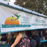 A banner for the Ovarian Cancer Alliance with a green ribbon, sun, and cloud, displayed at an outdoor event with people in the background.