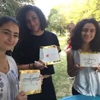 Three girls outdoors holding award certificates, smiling, with trees and sunlight in the background.