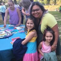 A woman with glasses and a yellow shirt smiling with two young girls in pink and blue dresses at an outdoor event, a table with craft supplies in front of them.