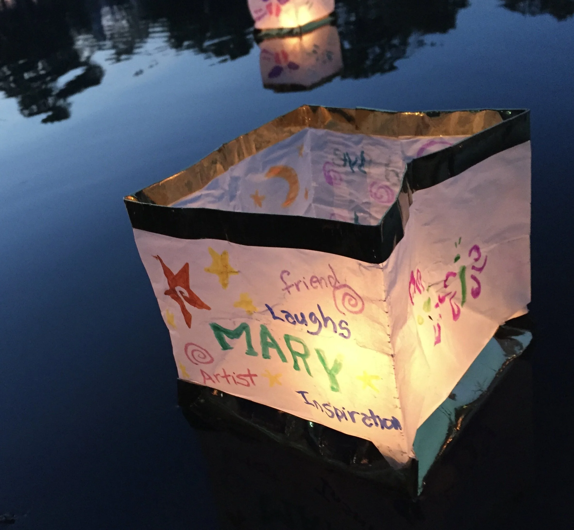 A paper lantern with colorful drawings and words like 'happy,' 'laughs,' 'friend,' 'artist,' and 'inspiration' floats on dark water during twilight.
