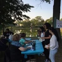 People gathered around a table outdoors by a lake, engaging in an activity, with trees and a sign visible in the background.