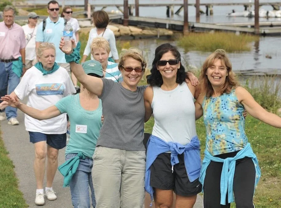 Group of women walking outdoors near water, smiling and celebrating with arms around each other, dressed casually for warm weather.