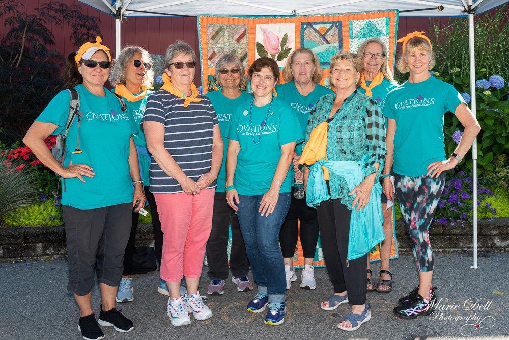 A group of nine women standing outdoors in front of a colorful quilt hanging under a canopy, all wearing matching teal t-shirts with the word 'Ovation' printed on them, some with yellow neckerchiefs and sunglasses, amid a garden with purple and pink flowers.