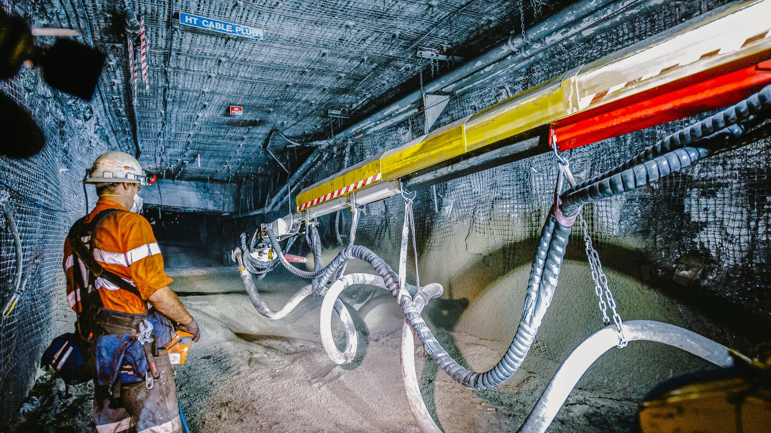 Worker in orange safety gear and helmet inside a tunnel with various pipes, hoses, and chains for construction or maintenance work.