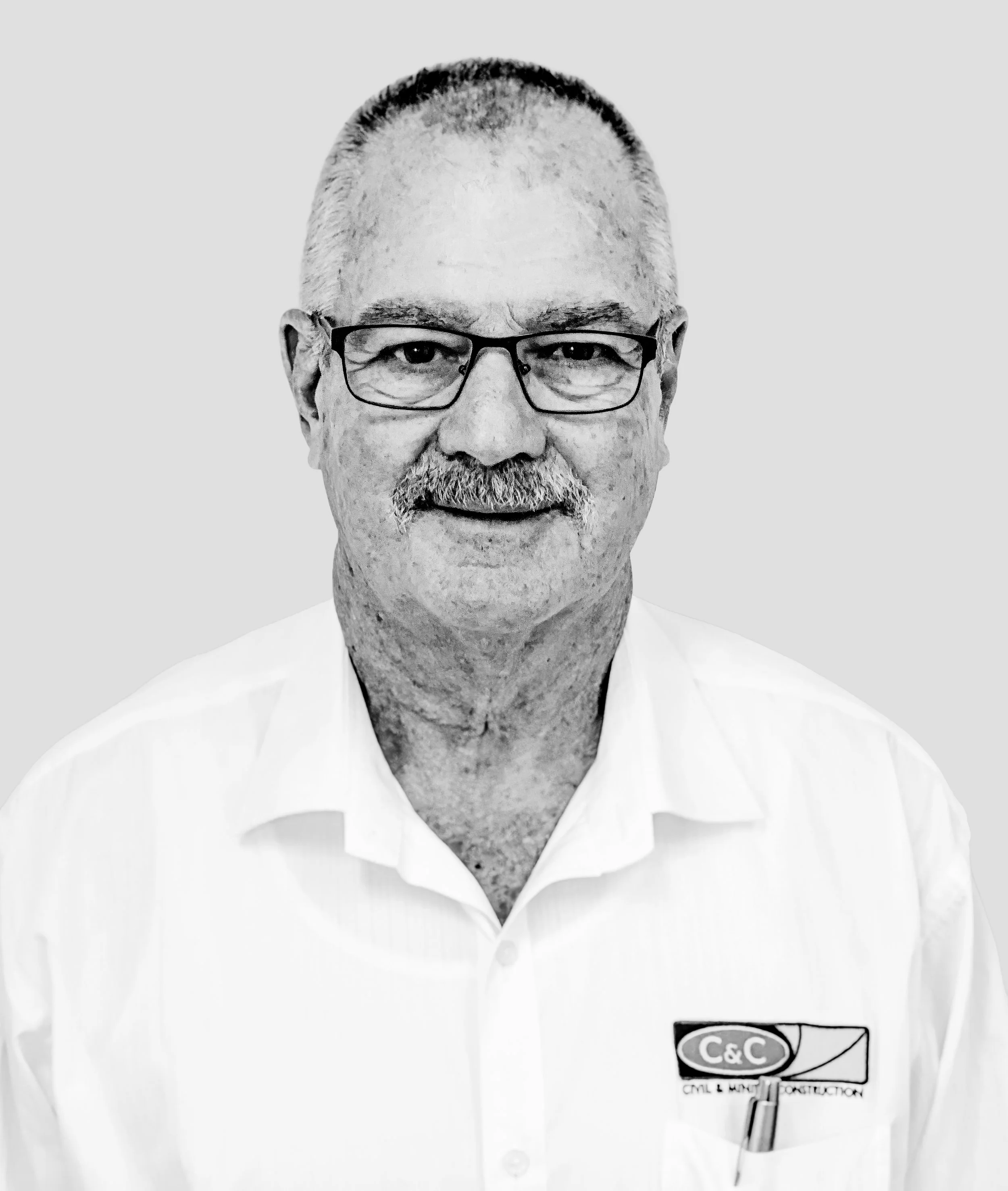 Black and white photograph of man's face, middle-aged, with glasses, short hair, beard, wearing a white shirt with a logo and text that reads 'C&C Civil & Mechanical Construction', against a plain light background.