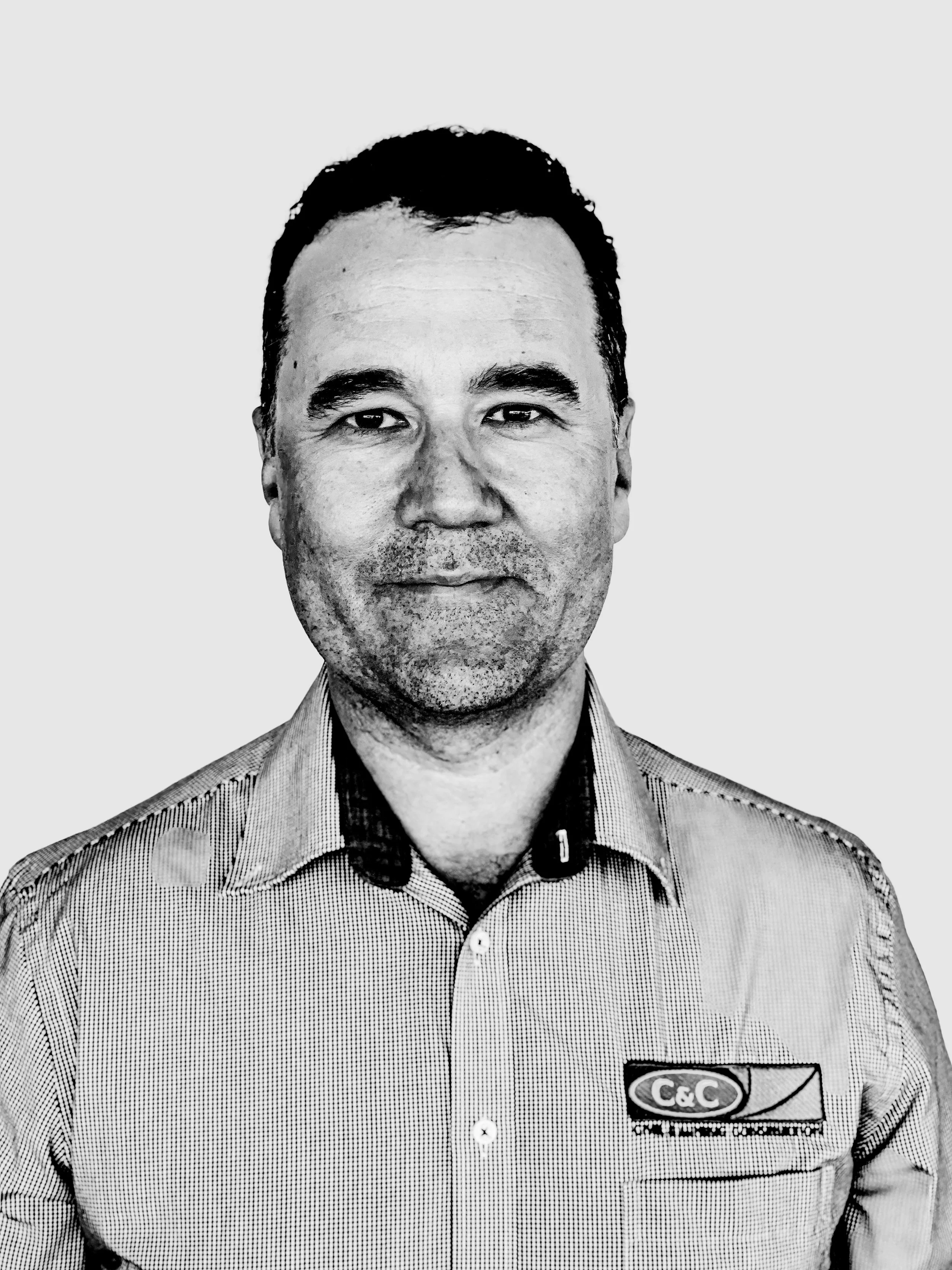 Black and white photo of a man wearing a checkered collared shirt with a logo on the pocket, smiling slightly, with a plain light background.