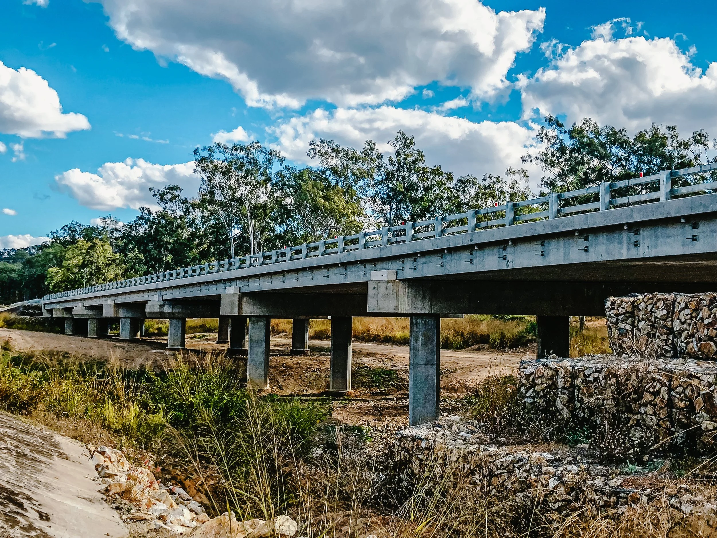 A concrete bridge over a dry, grassy landscape with some trees and a blue sky with clouds.