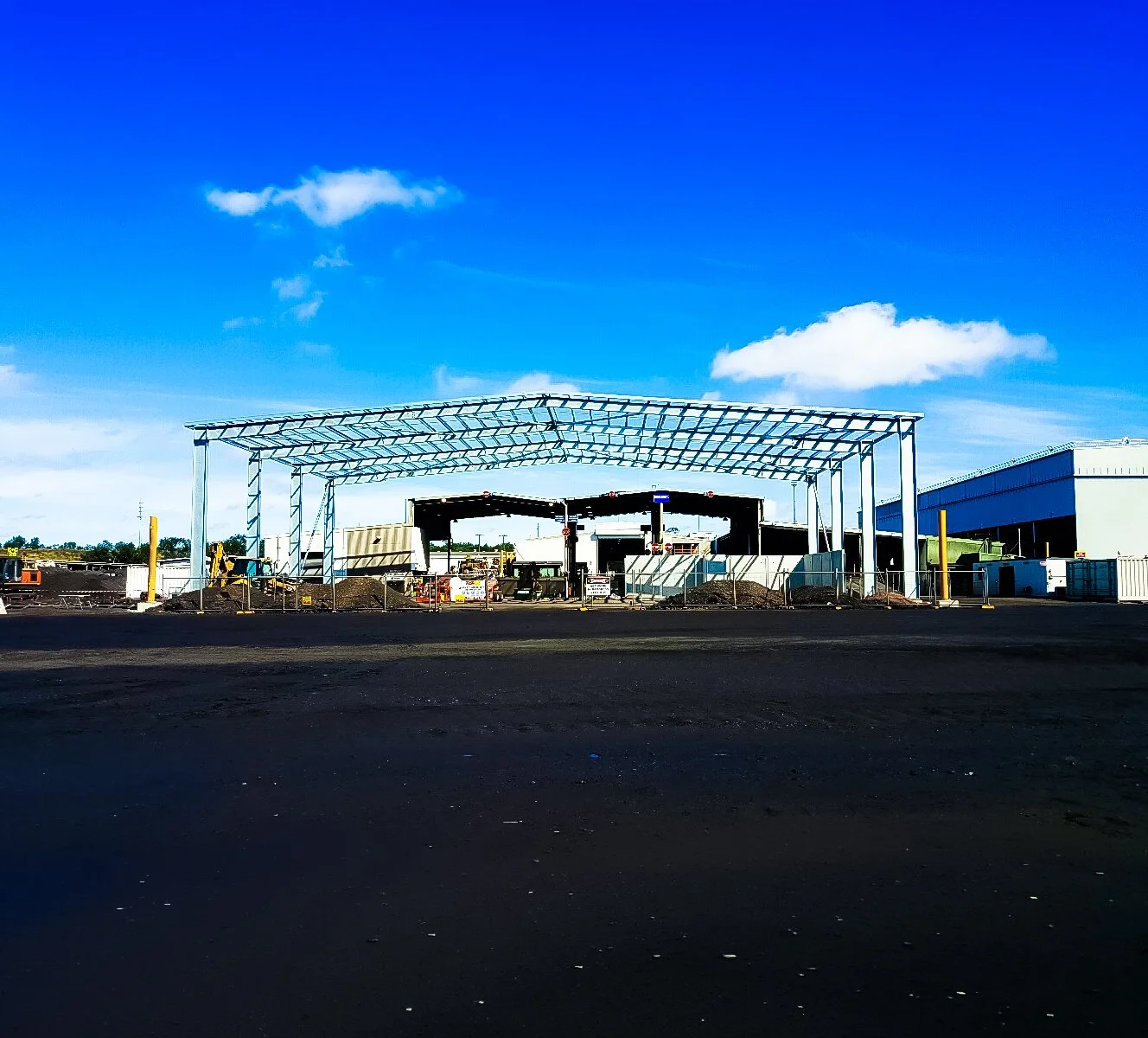 Construction site with a partially built metal framework, construction equipment, and piles of dirt under a blue sky with scattered clouds.
