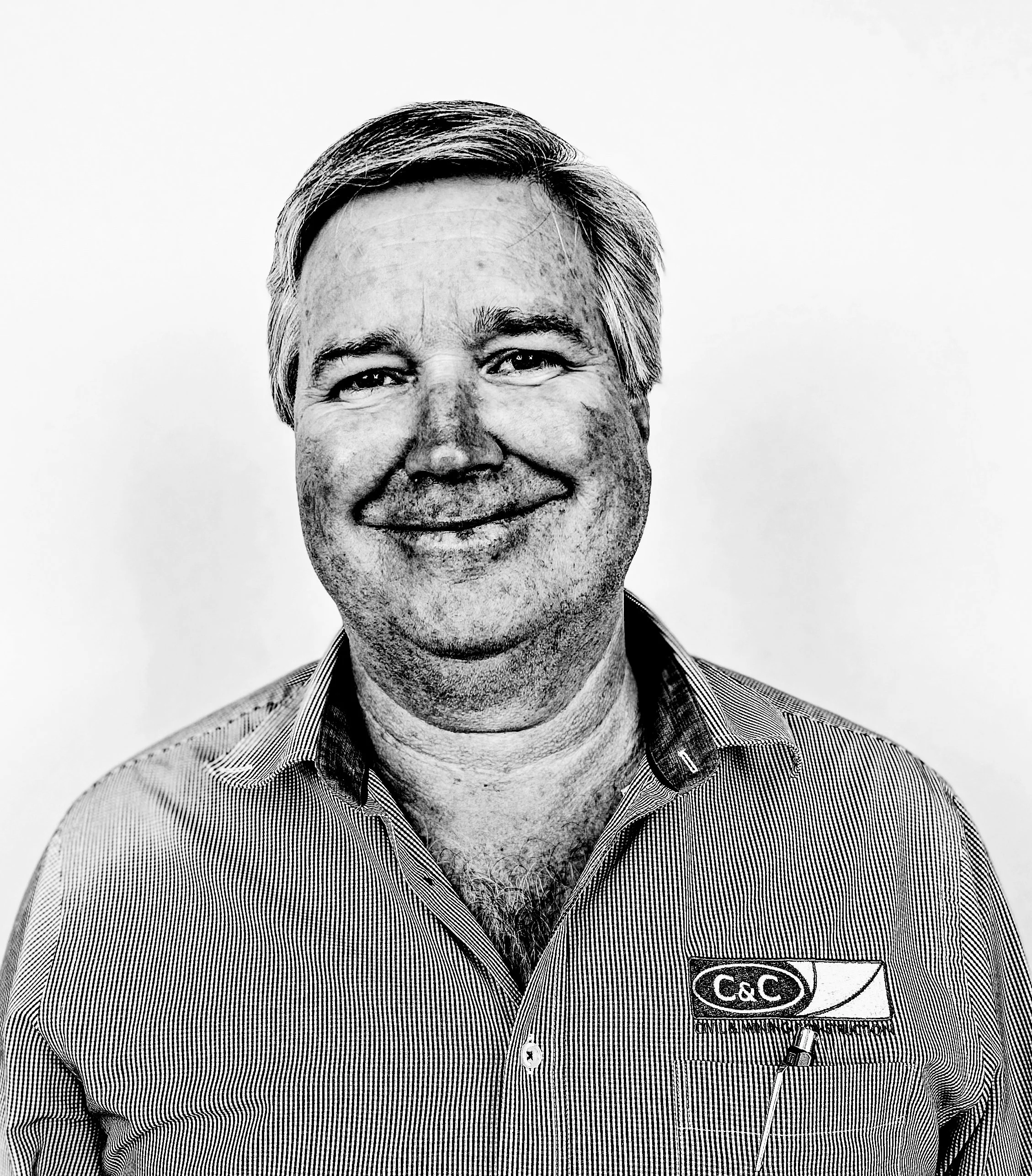 Close-up black-and-white photo of a smiling middle-aged man with short, graying hair, wearing a striped shirt with a logo that says "C&C" and a name tag. He's wearing a headset.