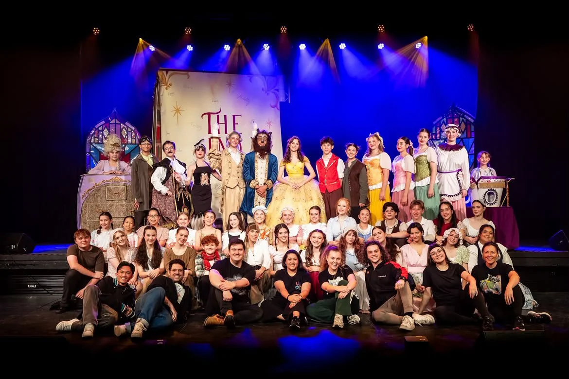 A large group of children and adults dressed in costumes from 'Beauty and the Beast' on stage, with a backdrop of a closed book that says 'The End' in a theatrical setting.