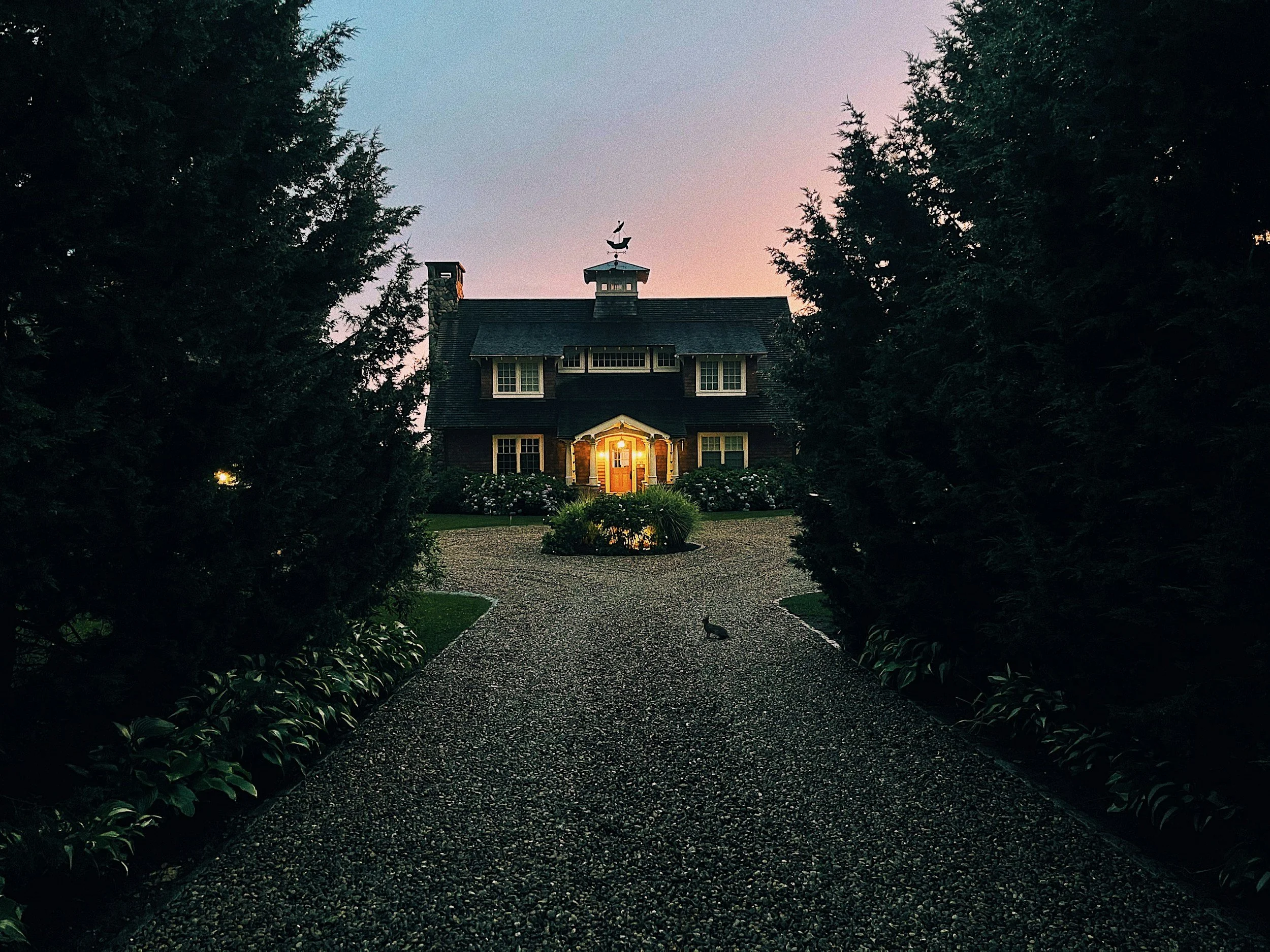 A house surrounded by trees at dusk, with a gravel pathway leading to the front door illuminated by a porch light.