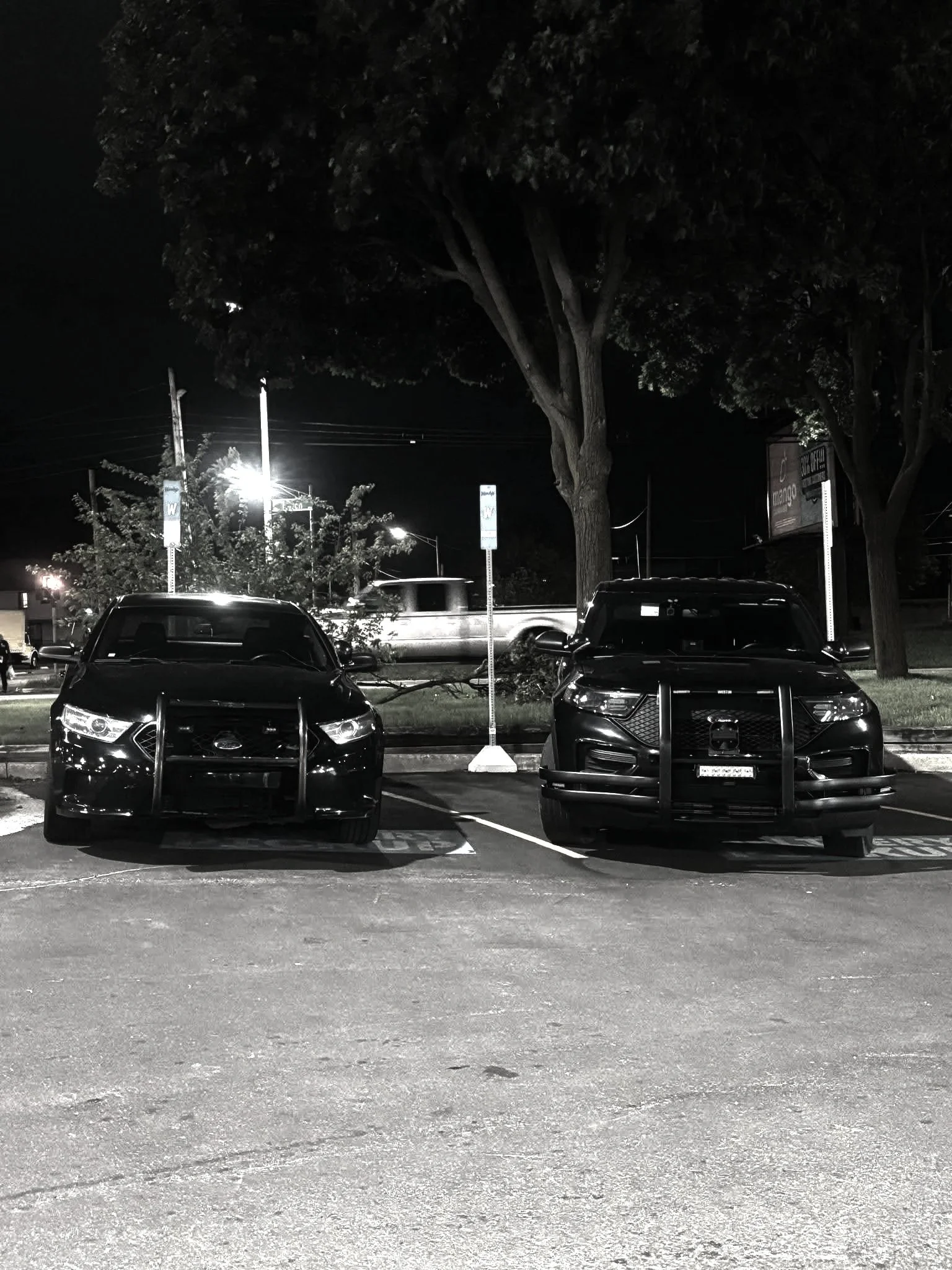 A black car and a dark SUV parked in a parking lot at night, with trees and streetlights in the background.