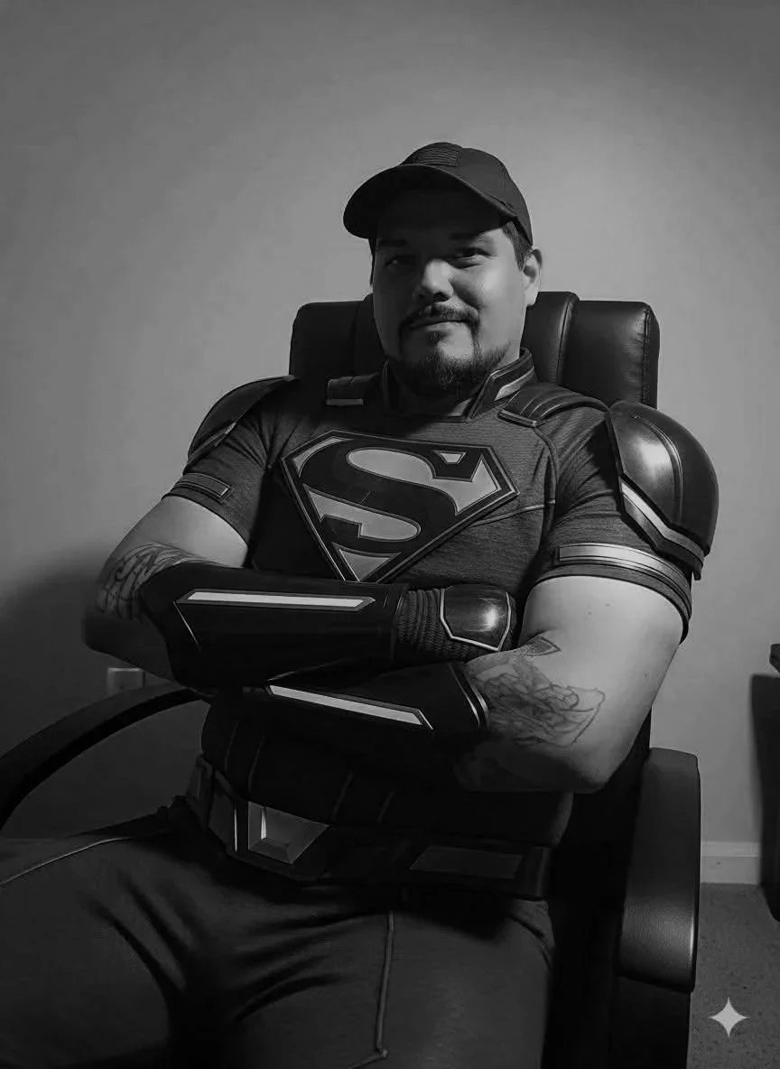 A man sitting in a chair wearing a Superman t-shirt, armor shoulder pads, a cap, and arm sleeves, with his arms crossed and smiling.