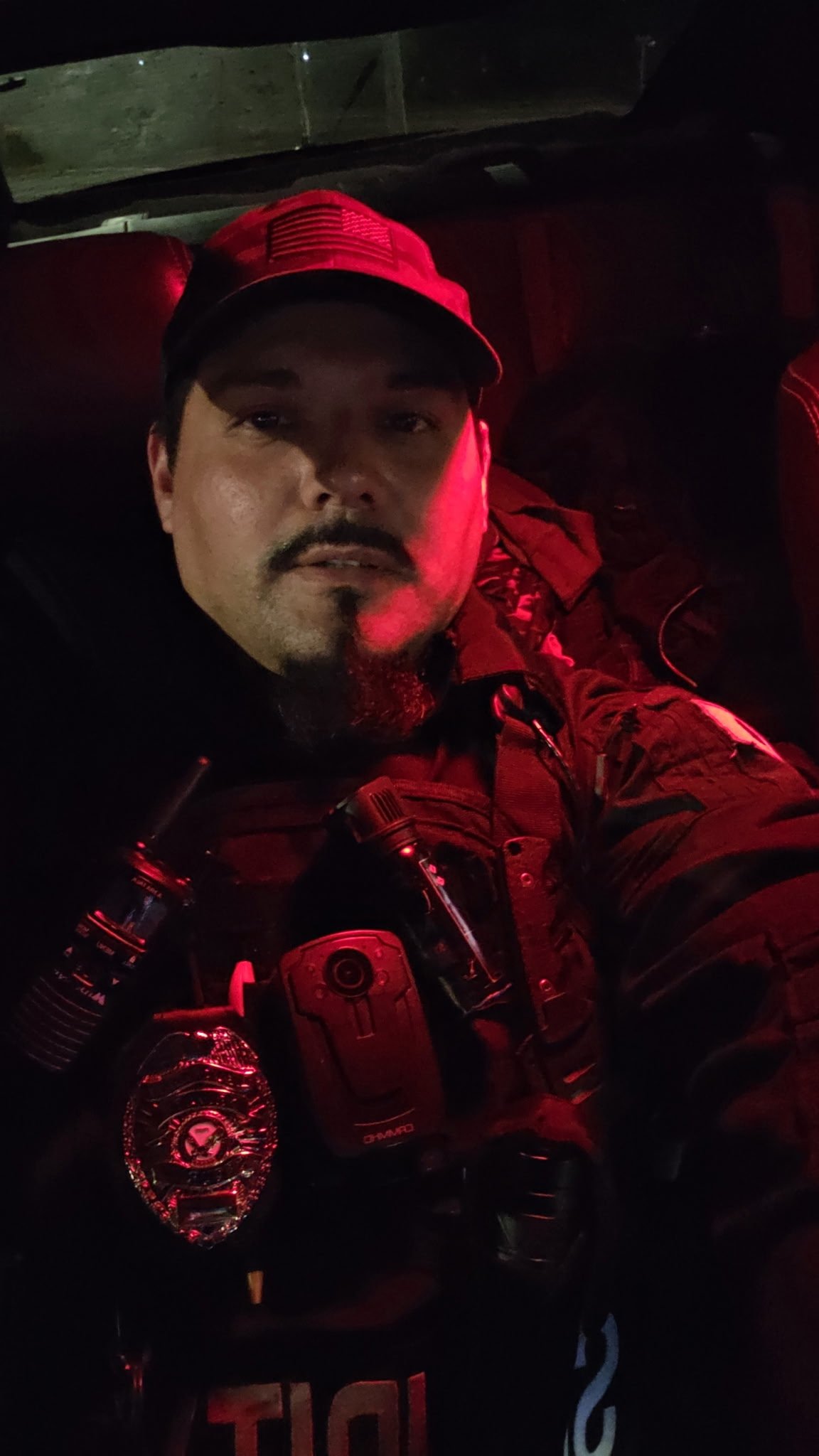 A man in a police uniform taking a selfie inside a vehicle at night. He is wearing a black cap with a badge, a tactical vest with equipment, and there is a police patch on his sleeve. Red light illuminates his face and the interior of the vehicle.