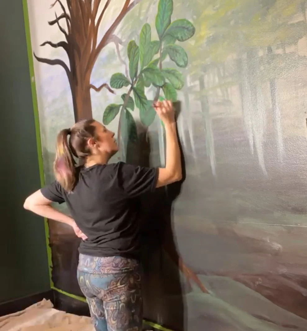 Alicia Maye painting a freehand forest mural. Green leaves and tree branches on dark wall.