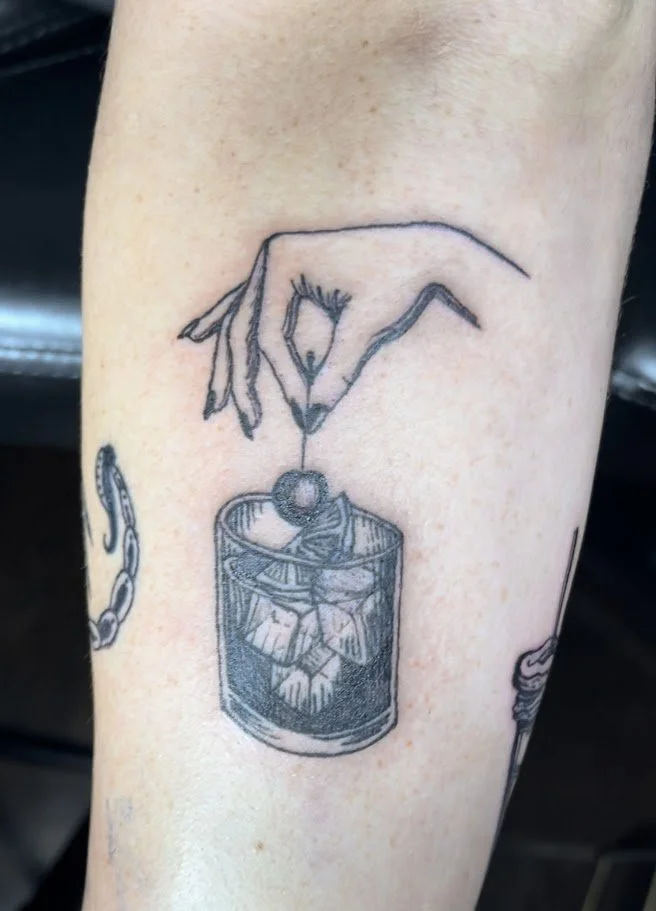 Tattoo of a hand holding a cherry above a glass with ice and mint leaves.