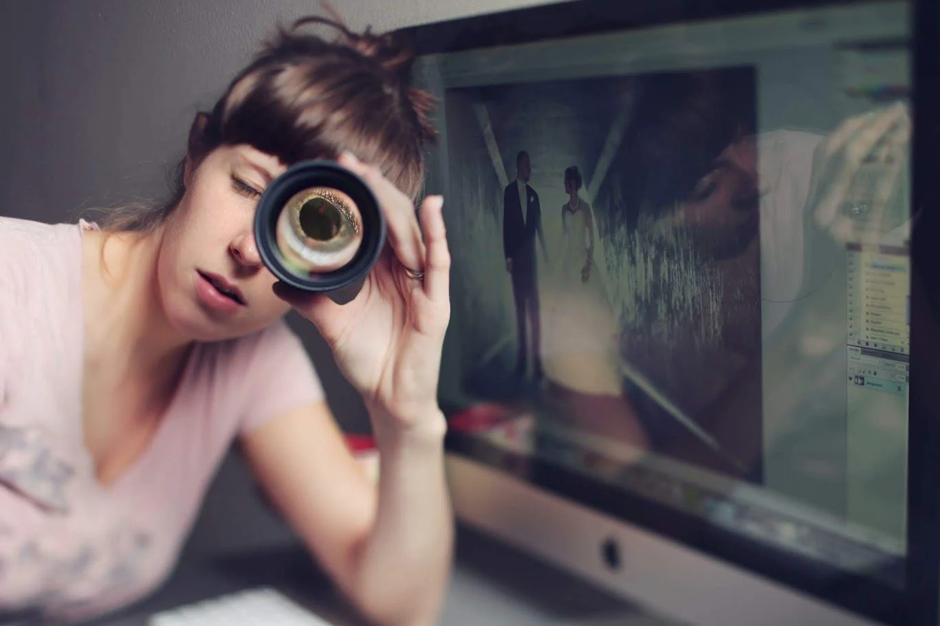 Alicia Maye holding a camera lens to her eye with wedding photo editing on screen behind her. Wedding photographer portrait.