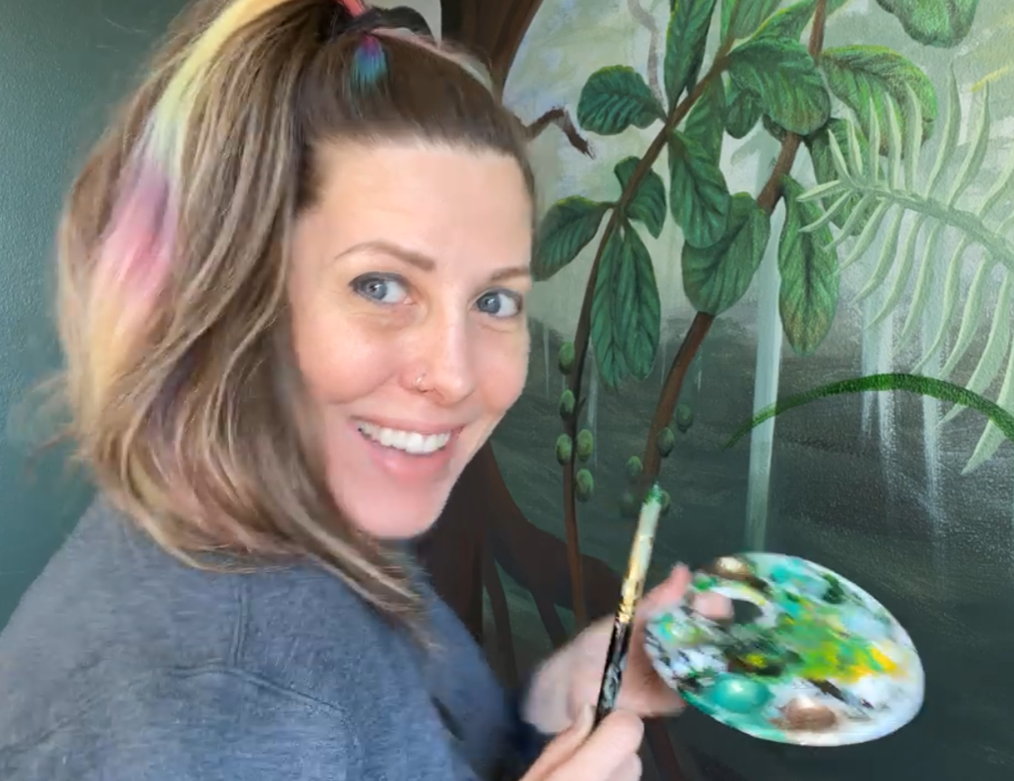 Alicia Maye holding a paintbrush and palette, smiling in front of a tropical leaf painting in progress.
