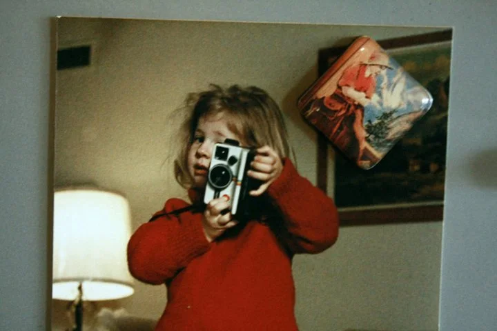 Alicia Maye as a toddler holding a camera. Red sweater, mirror selfie. The beginning of a lifelong artist.
