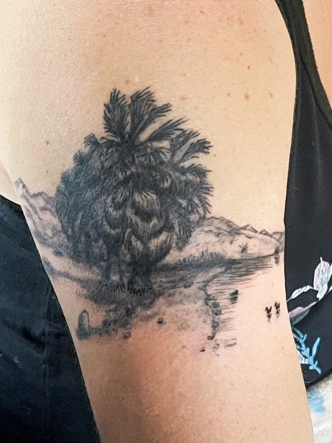 Tattoo of a palm tree, mountain range, and water scene on the upper arm.