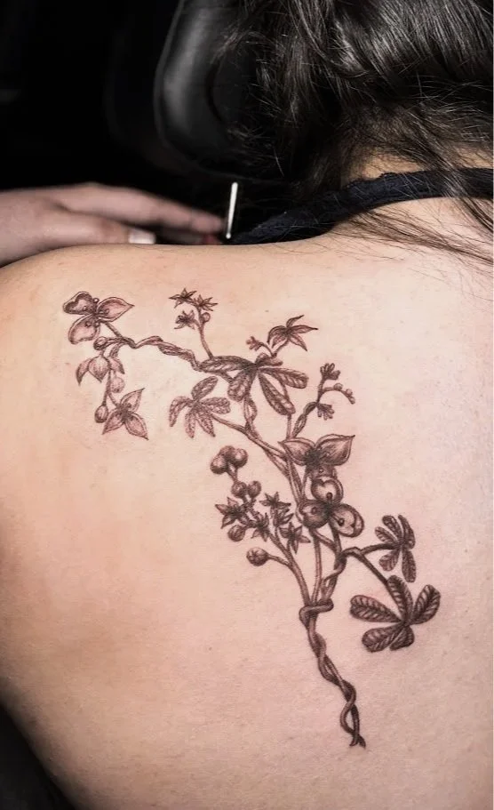A close-up of a woman's shoulder with a freshly inked, intricate botanical tattoo featuring intertwined vines, leaves, and small flowers.