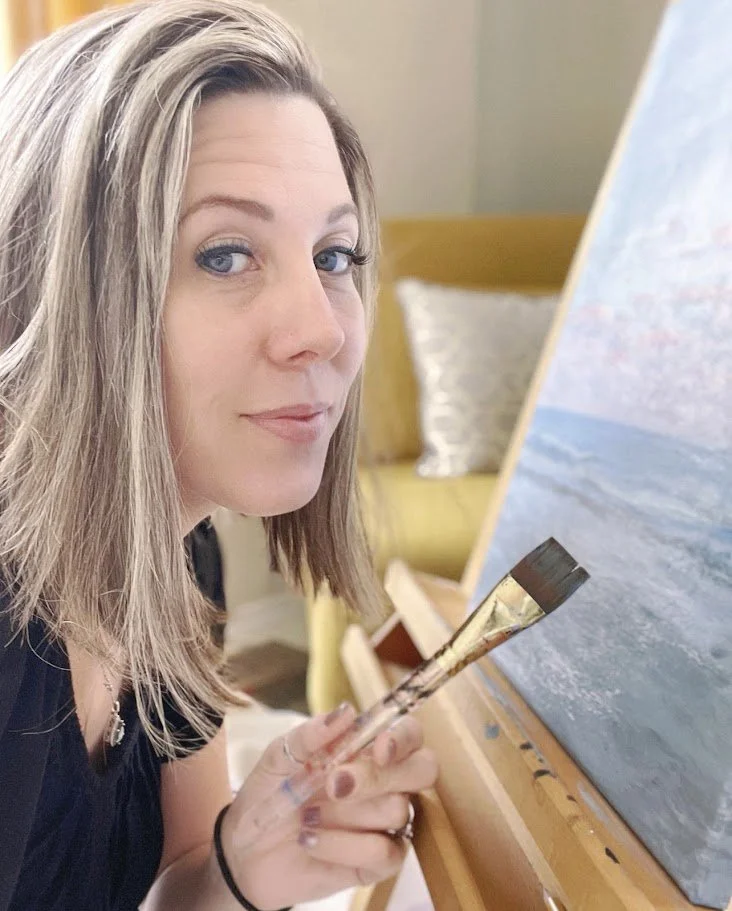 Alicia Maye painting a landscape on easel, holding palette and brush. Studio process shot.