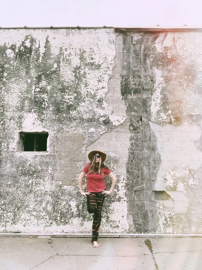 Alicia Maye artist portrait. Red shirt, tan hat, barefoot against a weathered wall.