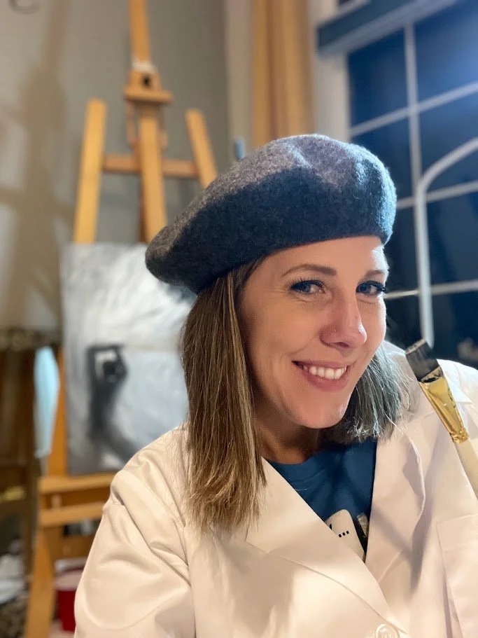 Alicia Maye in her art studio, smiling with easel and painting in background.