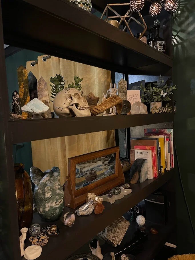 A black bookshelf filled with various decorative items, including stones, crystals, books, a skull sculpture, and small plants, with a wooden cabinet and yellow curtain in the background.