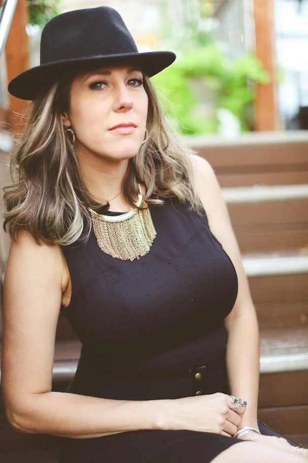 Artist portrait of Alicia Maye. Black hat, gold necklace, outdoor stairs.