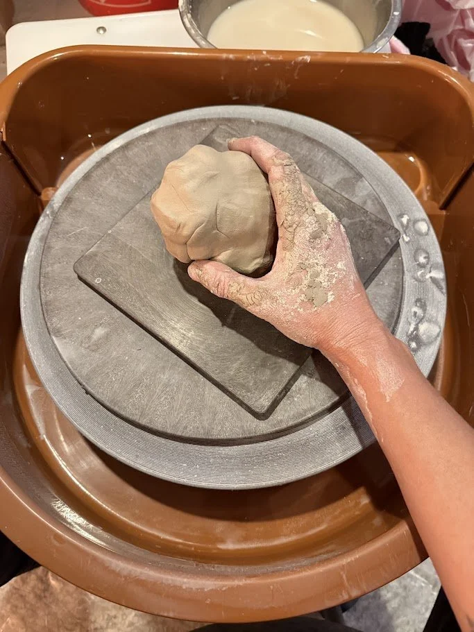A person shaping a ball of clay with their hand on a pottery wheel. The person's hand and arm are covered in clay dust, and the pottery wheel is set inside a brown basin. In the background, there is a bowl with a creamy liquid.