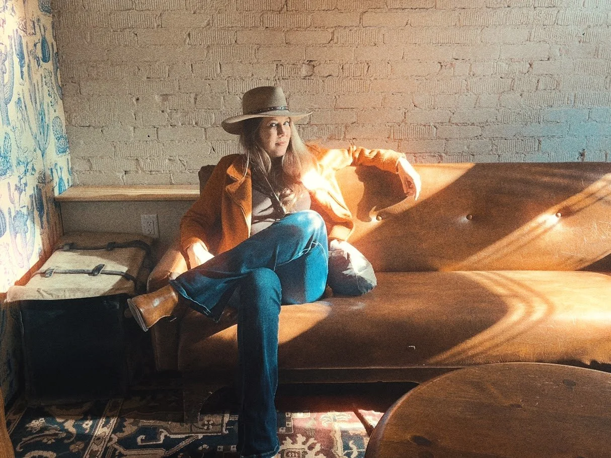 Alicia Maye artist portrait. Wide brim hat, orange blazer, brick wall background.