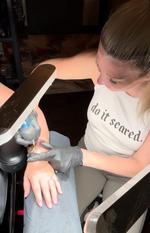A woman getting a tattoo on her hand at a tattoo parlor, with a tattoo artist working on her.