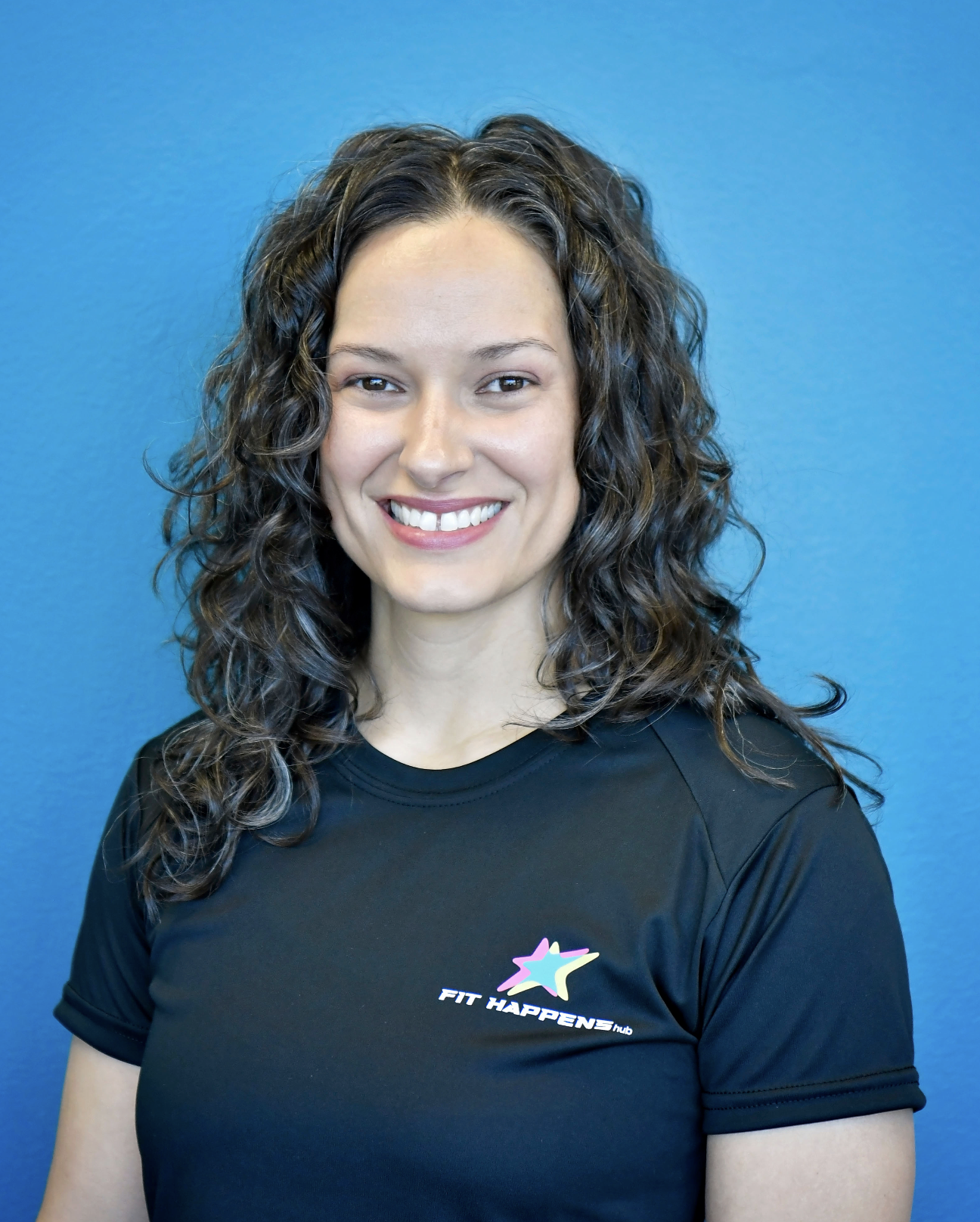 A woman with curly hair smiling while lifting a barbell in a gym setting.