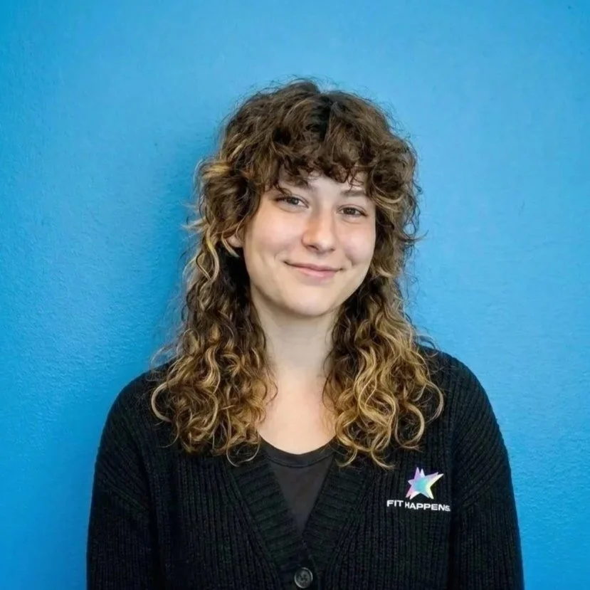 A woman with curly brown hair smiling in an office setting with a plant and motivational poster in the background.