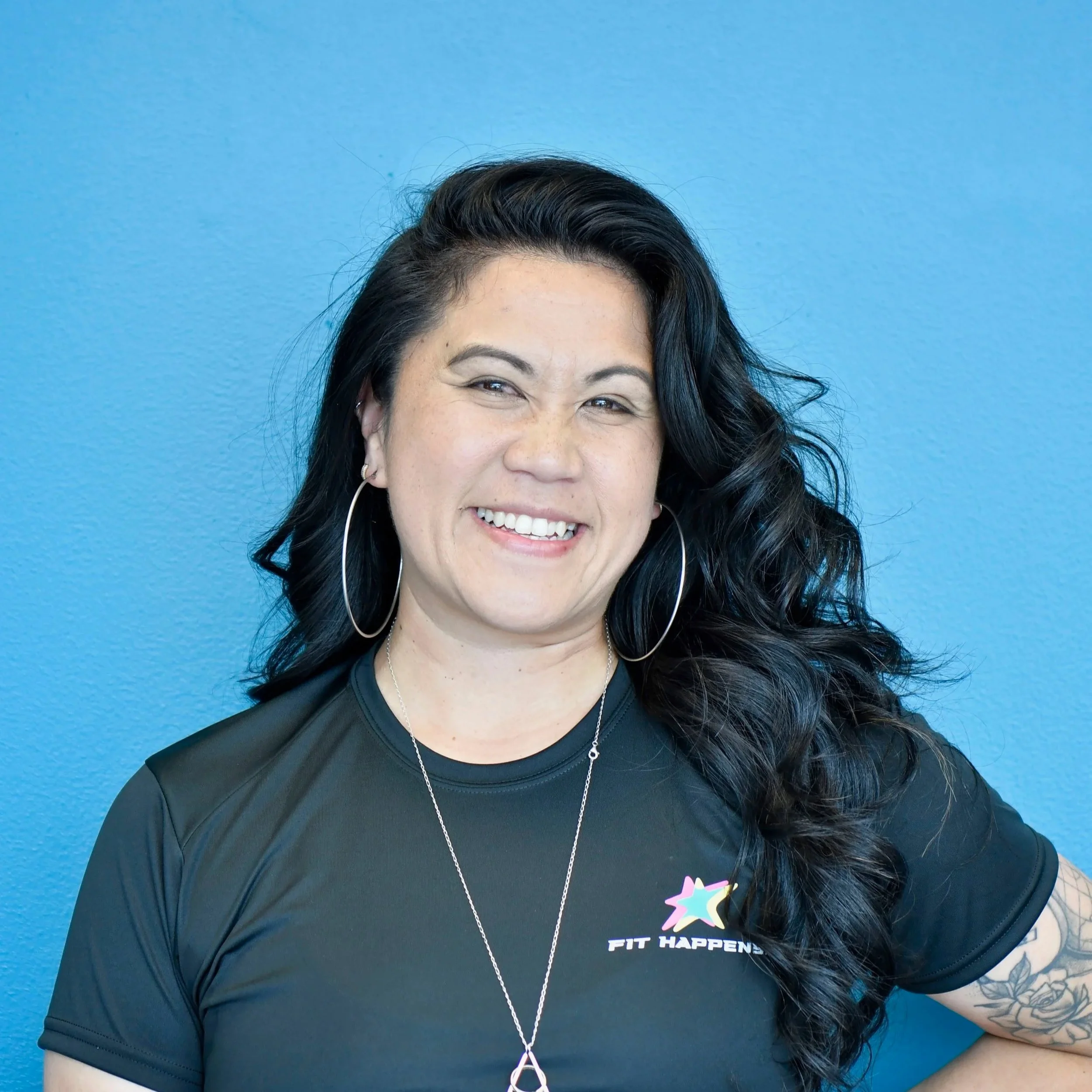 A woman with long dark hair and tattoos, winking and smiling in front of a dark background.