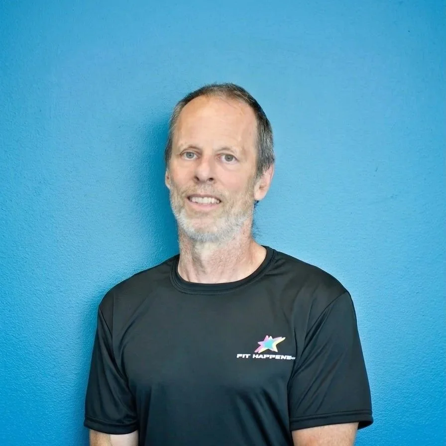 A middle-aged man with a beard and short hair smiling in a gym with black brick walls and dumbbells in the background.