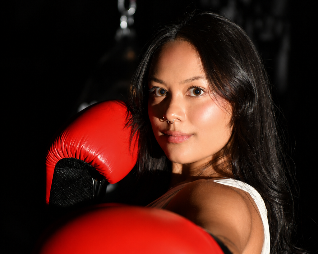 A woman with dark hair, blue eyes, and a septum piercing wearing red boxing gloves, standing in a boxing stance in a dark gym.
