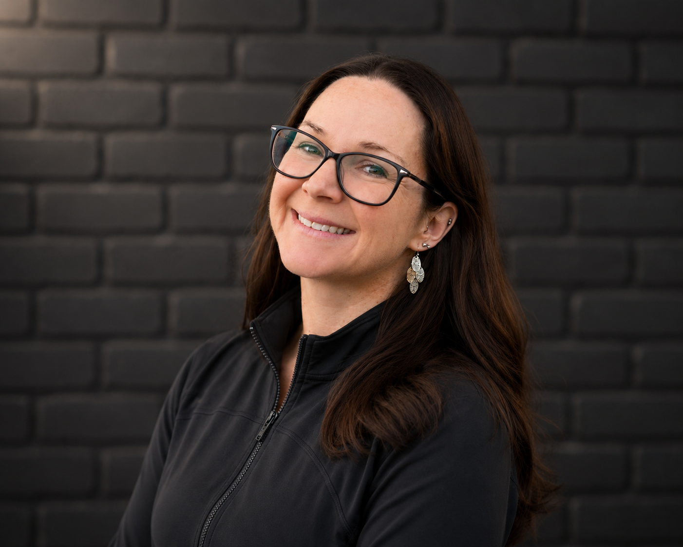 A woman with long dark hair and glasses smiling in front of a black brick wall.