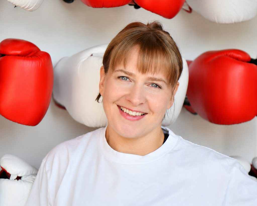 A woman with auburn hair and a white shirt smiling, surrounded by red and white boxing gloves.