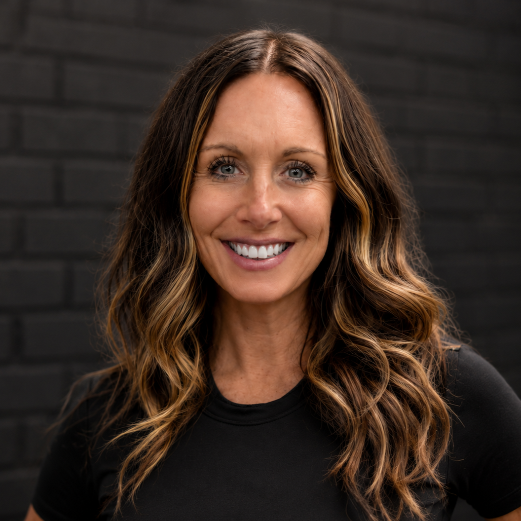 A woman with long wavy brown hair with blonde highlights, smiling and wearing a black t-shirt, standing against a dark brick wall.