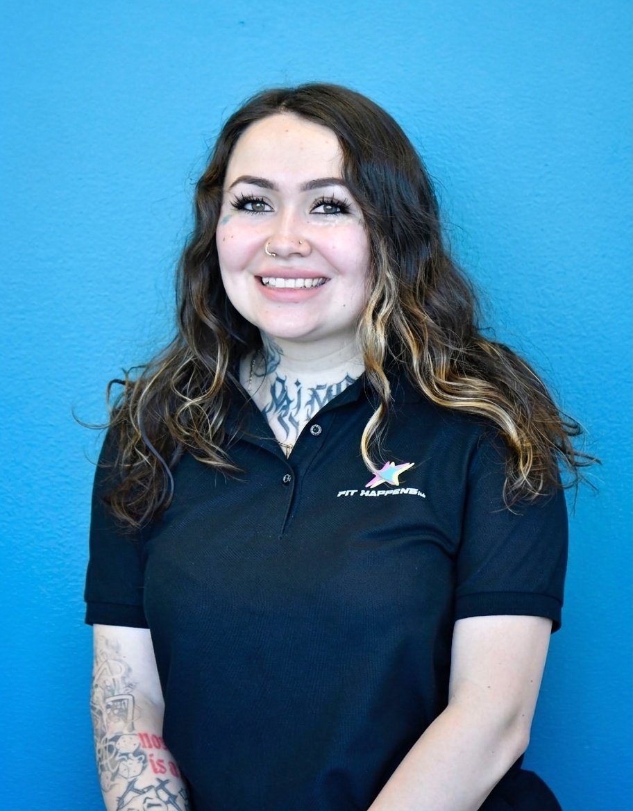 A smiling woman with curly brown hair and tattoos on her neck and arms, sitting at a reception desk with a welcome sign in front. Behind her are large windows, plants, kettlebells, and a sign on the wall that says 'Stronger Together'.