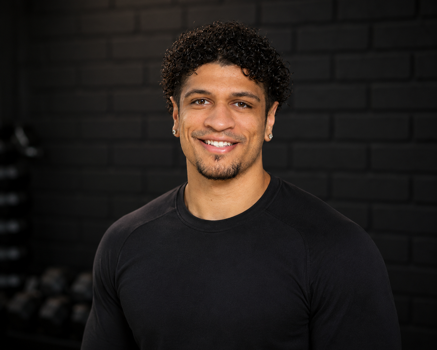A smiling man with curly dark hair, earrings, and a goatee wearing a black long-sleeve shirt, standing against a dark brick wall background.