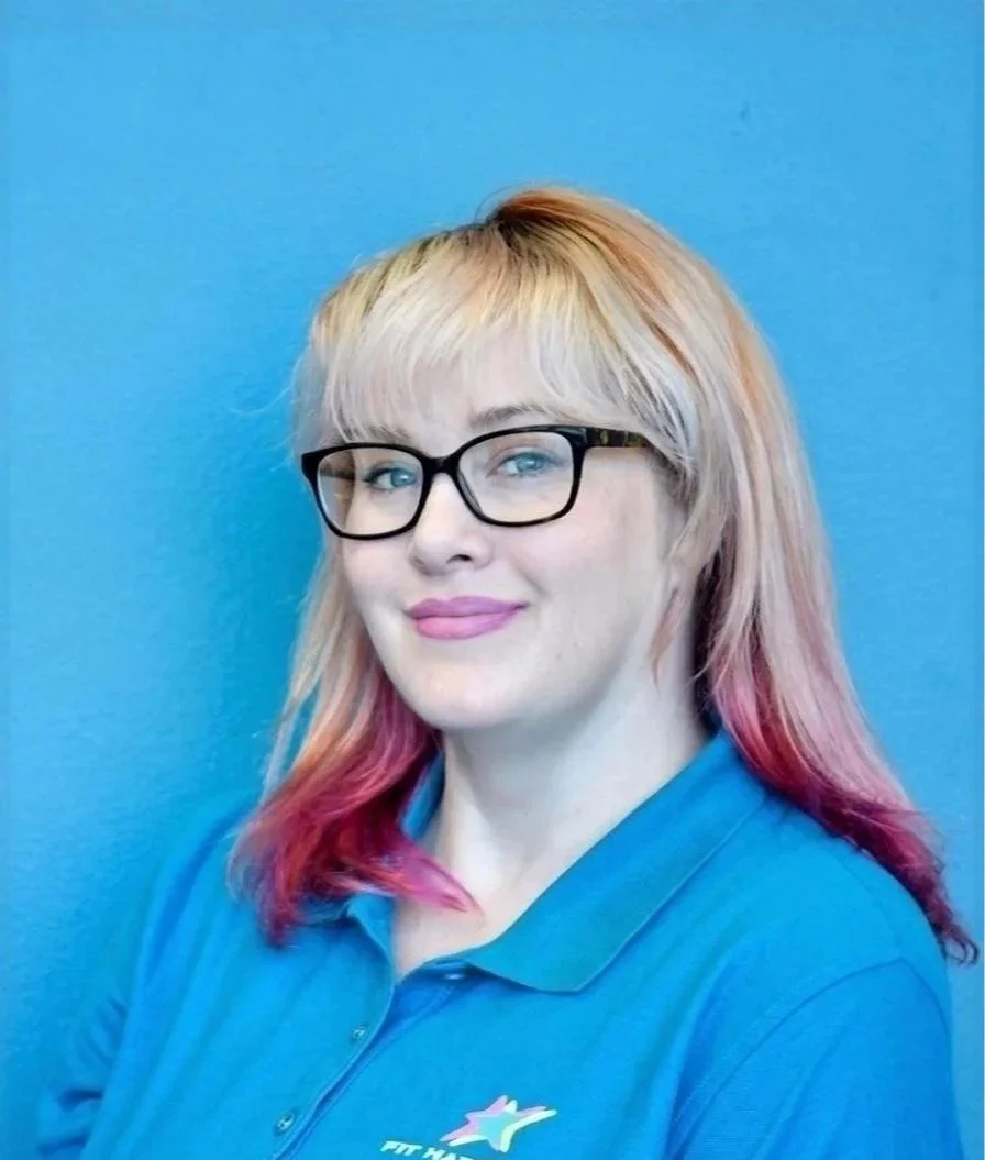 A woman with blonde hair, glasses, and a black top sitting at a reception desk with a sign that reads 'Welcome, we're glad you're here!' behind her is a black wall with the words 'Stronger Together, Fit Happen' and a shelf with black items, a window, and a potted plant.